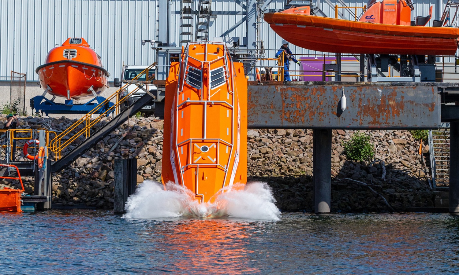 Un bote salvavidas de color naranja brillante se lanza al agua desde un muelle, creando un chapoteo. Al fondo se ven otros botes salvavidas y estructuras industriales.