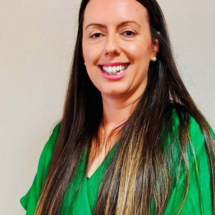 A smiling woman with long dark hair wearing a green blouse stands against a plain background.