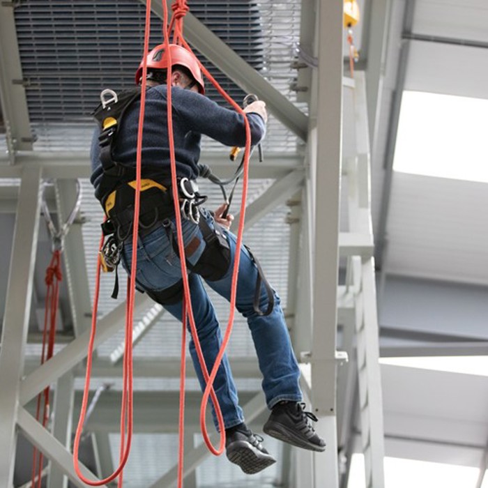 A person wearing safety gear and a red helmet is suspended by ropes, climbing on a metal structure inside an industrial facility.
