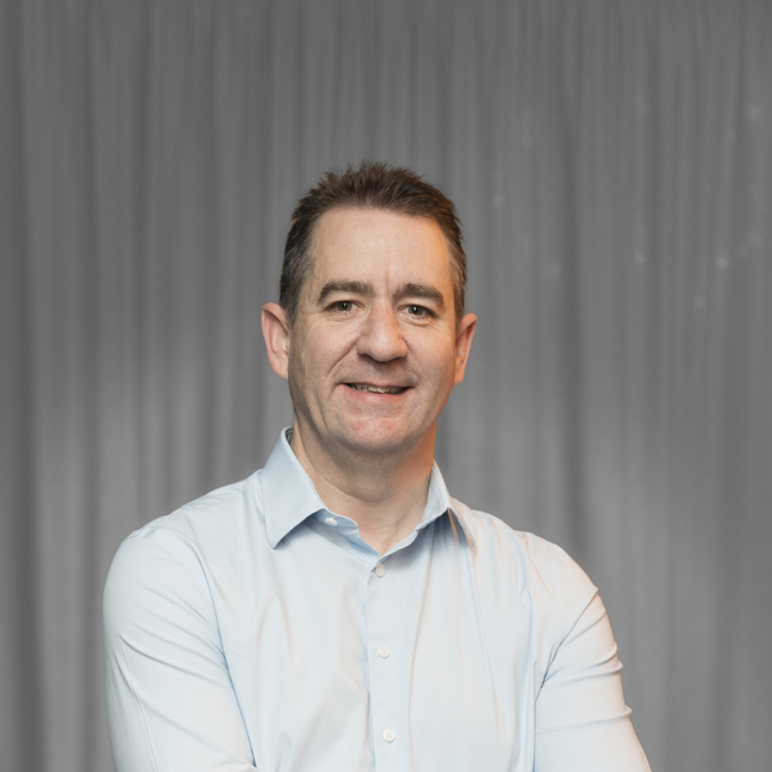 رجل يرتدي قميصًا أزرق فاتحًا ويقف واضعًا ذراعيه متشابكًا ومبتسمًا أمام خلفية ستارة رمادية.