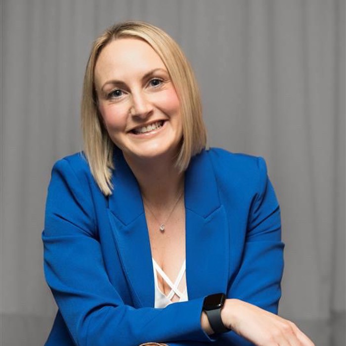 شخص يرتدي بدلة زرقاء وساعة ذكية ويبتسم وهو جالس واضعًا ذراعيه على ركبتيه، وفي الخلفية ستارة رمادية.