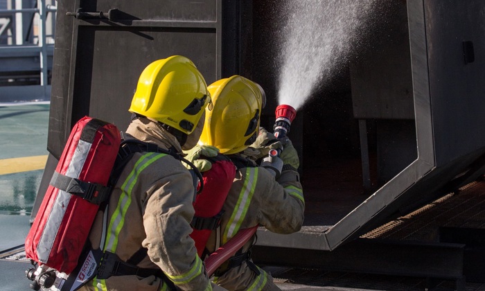 Bomberos con cascos amarillos y equipo de protección rocían agua en una estructura en llamas durante un ejercicio de entrenamiento.