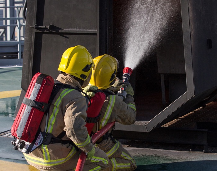 Des pompiers portant des casques jaunes et des équipements de protection pulvérisent de l'eau sur une structure en feu lors d'un exercice d'entraînement.