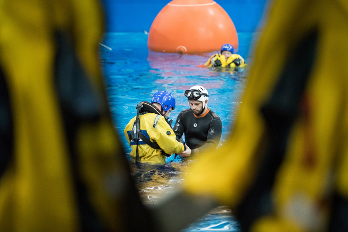 Dois indivíduos com equipamento para actividades aquáticas são vistos no centro de uma piscina, rodeados por outros em fatos amarelos, com uma grande boia cor de laranja ao fundo.