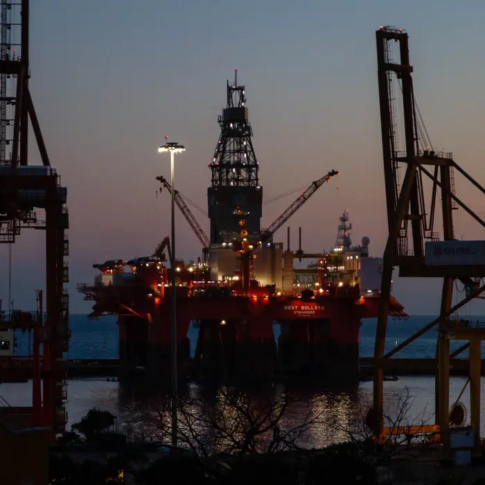 Una plataforma petrolífera en alta mar iluminada al atardecer, rodeada por las siluetas de grandes grúas y estructuras industriales, con el mar de fondo.