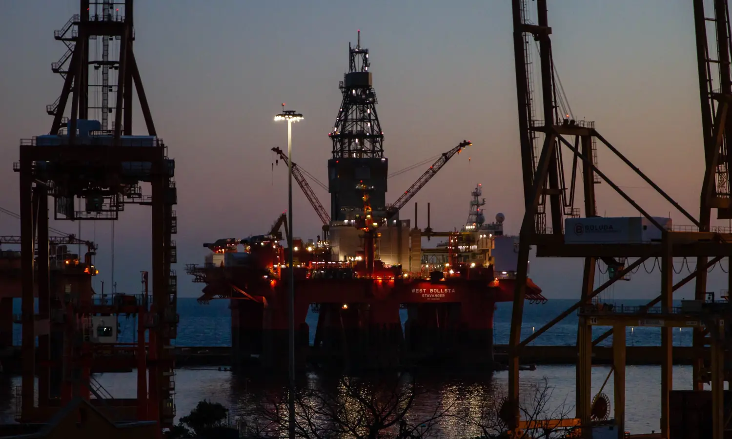Uma plataforma petrolífera offshore iluminada ao anoitecer, rodeada pelas silhuetas de grandes gruas e estruturas industriais, com o mar ao fundo.