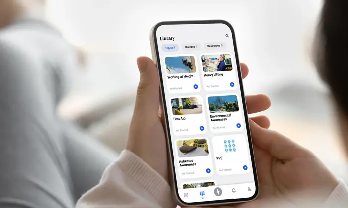 Pessoa que utiliza uma aplicação para smartphone para navegar numa biblioteca de cursos, incluindo "Trabalho em altura", "Primeiros socorros", "Sensibilização para o amianto", entre outros, com opções categorizadas em vários tópicos e sectores.