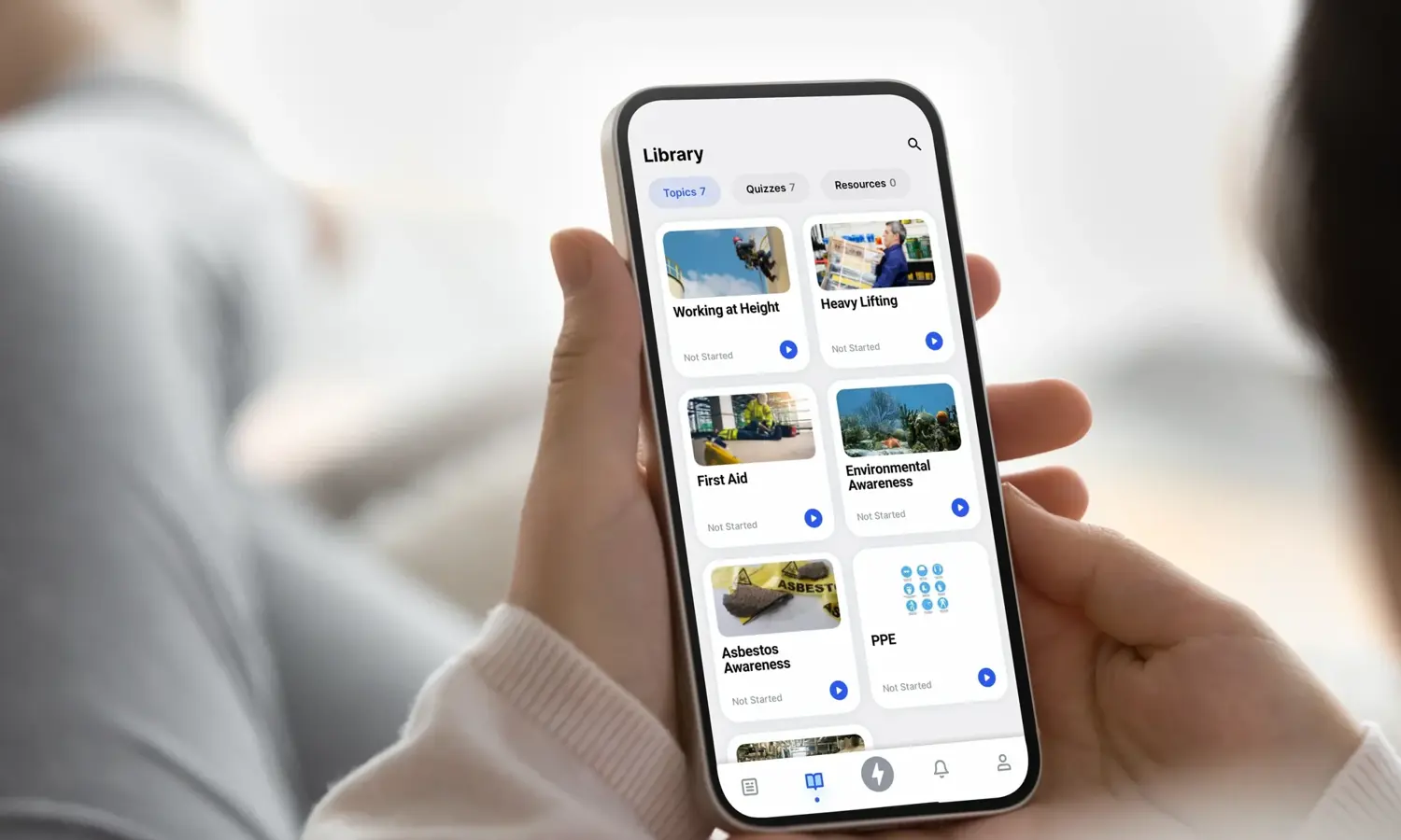 Persona que utiliza una aplicación de smartphone para navegar por una biblioteca de cursos que incluye "Trabajo en altura", "Primeros auxilios" y "Concienciación sobre el amianto", entre otros, con opciones clasificadas por temas y sectores.