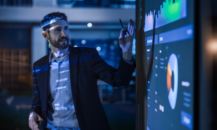 رجل يرتدي بدلة عمل يشير إلى شاشة تفاعلية كبيرة تعرض رسوماً بيانية ومخططات في مكتب حديث.