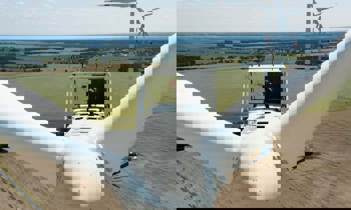 Un técnico está de pie encima de un aerogenerador en una zona rural, realizando tareas de mantenimiento. El paisaje circundante está formado por extensos campos y árboles lejanos.