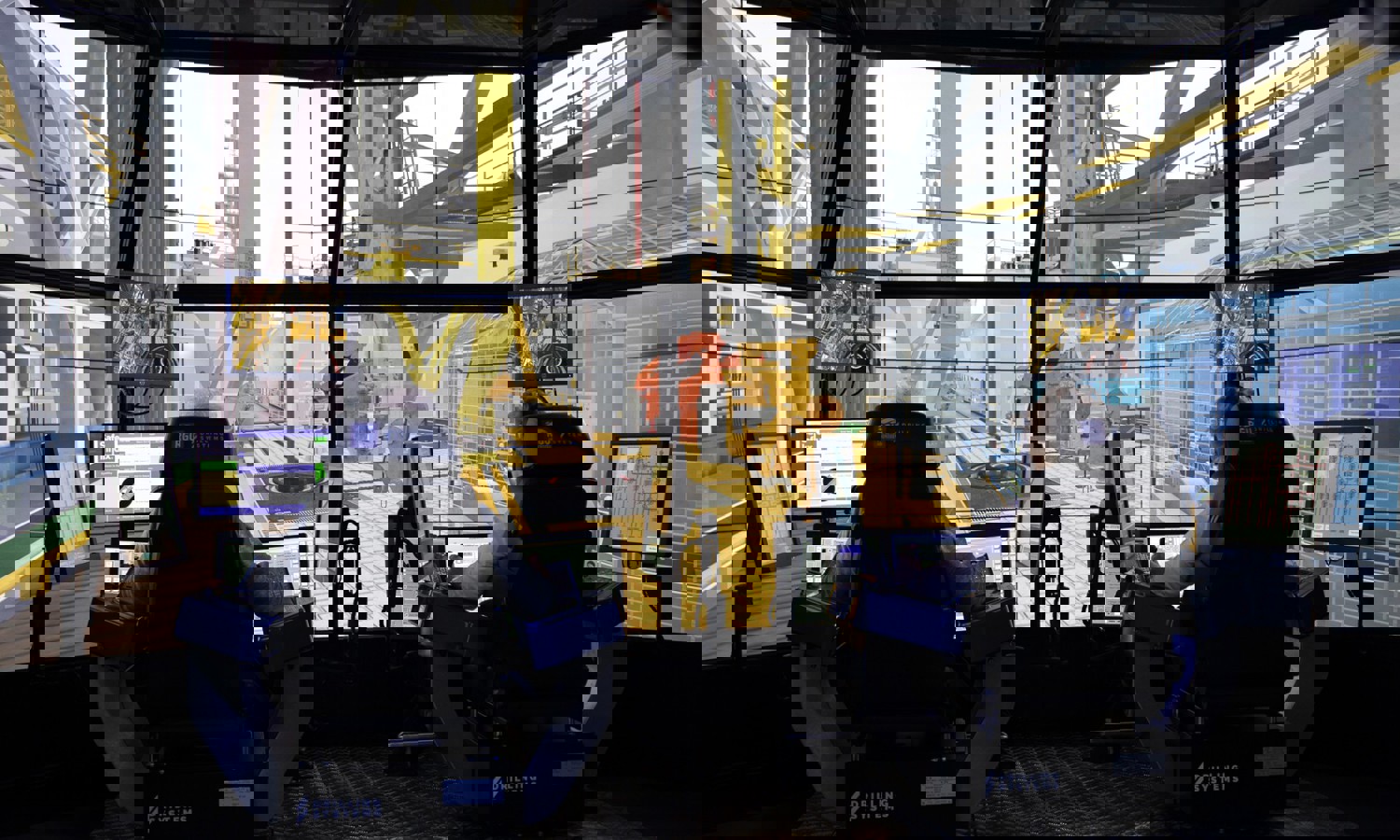 يجلس شخصان في محطتي تحكم مزودتين بشاشات متعددة، ويقومان بتشغيل جهاز محاكاة حفر عالي التقنية. شاشات كبيرة تحاكي بيئة الحفر البحرية من حولهم.