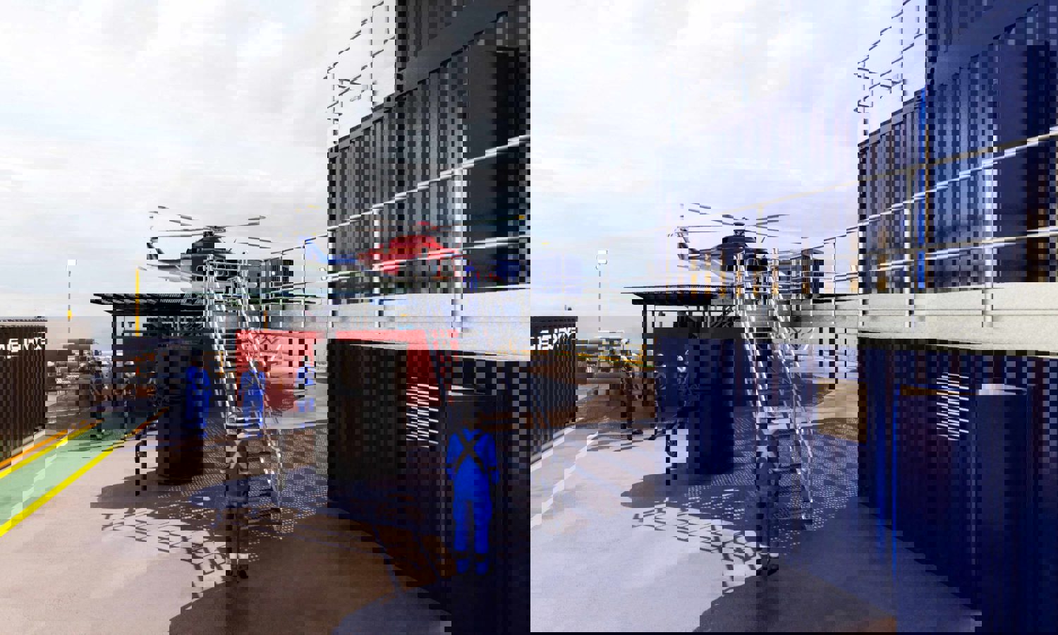 Helicóptero sobre una plataforma con trabajadores de uniforme azul cerca, situado junto a contenedores metálicos.