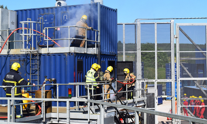 Bombeiros com uniformes amarelos e pretos realizam um exercício de treino, utilizando mangueiras e equipamento numa estrutura azul com vários níveis. O fumo é visível nos pisos superiores.