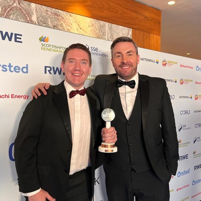 Two men in tuxedos stand on a red carpet. One holds an award trophy. The backdrop features logos of various sponsors.