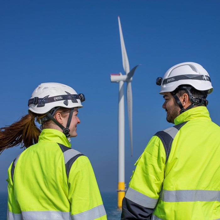 Two workers in high-visibility jackets and helmets, recently engaged in training, stand near an offshore wind turbine against a clear blue sky.