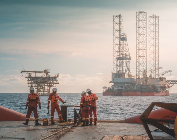 Cuatro trabajadores con equipo de seguridad en una plataforma en alta mar, perfeccionando sus habilidades mediante formación práctica mientras realizan tareas, con dos plataformas petrolíferas visibles al fondo sobre el mar.