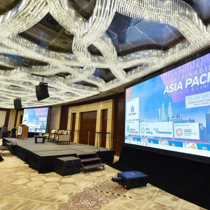 A conference room with attendees facing a stage and large screen displaying details of the "Asia Pacific Conference & Exhibition." The event is scheduled for 23-24 May 2023 in Kuala Lumpur.