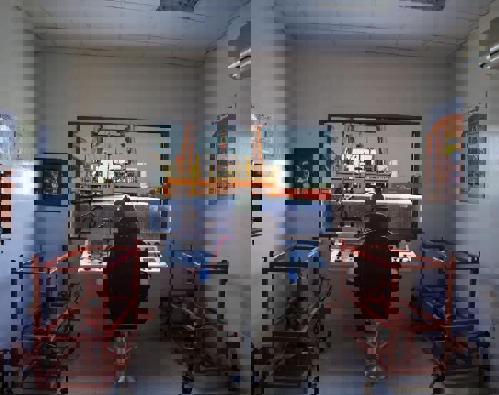 غرفة تحكم من الداخل بها شخص جالس على وحدة تحكم، يراقب رافعة ترفع حاوية في الخارج. إطارات معدات حمراء تحيط بالغرفة، وملصقات إعلامية على الجدران.