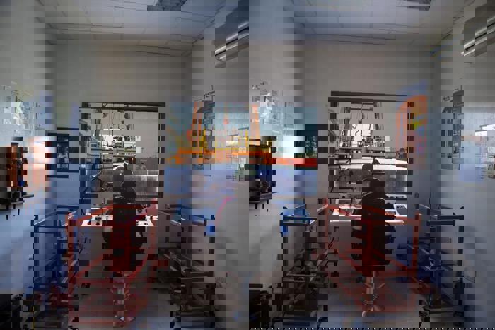 غرفة تحكم من الداخل بها شخص جالس على وحدة تحكم، يراقب رافعة ترفع حاوية في الخارج. إطارات معدات حمراء تحيط بالغرفة، وملصقات إعلامية على الجدران.