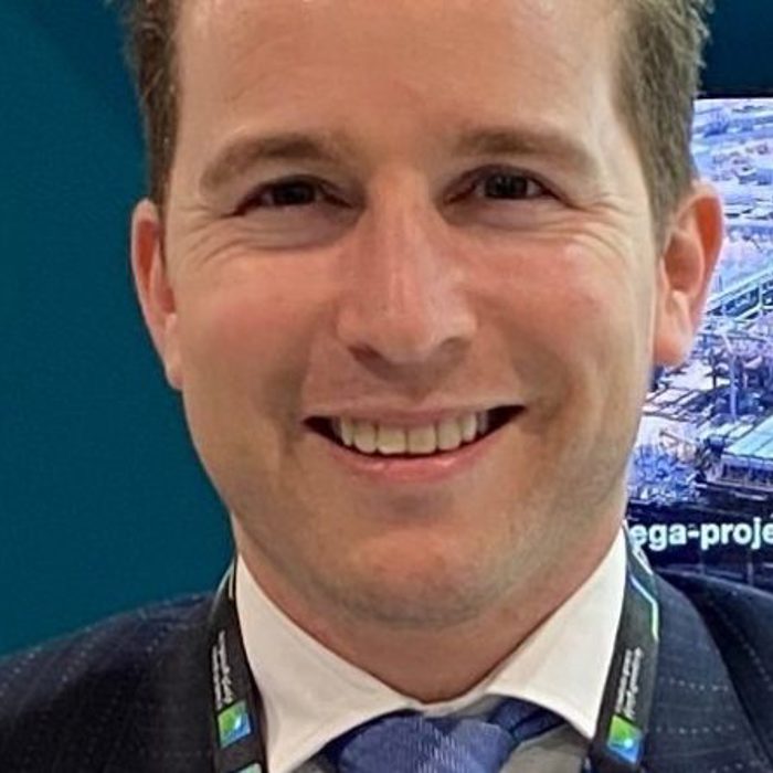 A smiling person in formal attire, with a lanyard around their neck, positioned in front of a screen displaying an industrial scene.