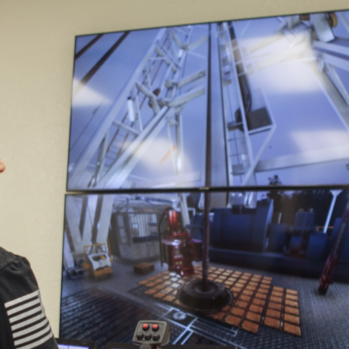 A woman operates a control panel while watching a large screen displaying machinery in an industrial setting.