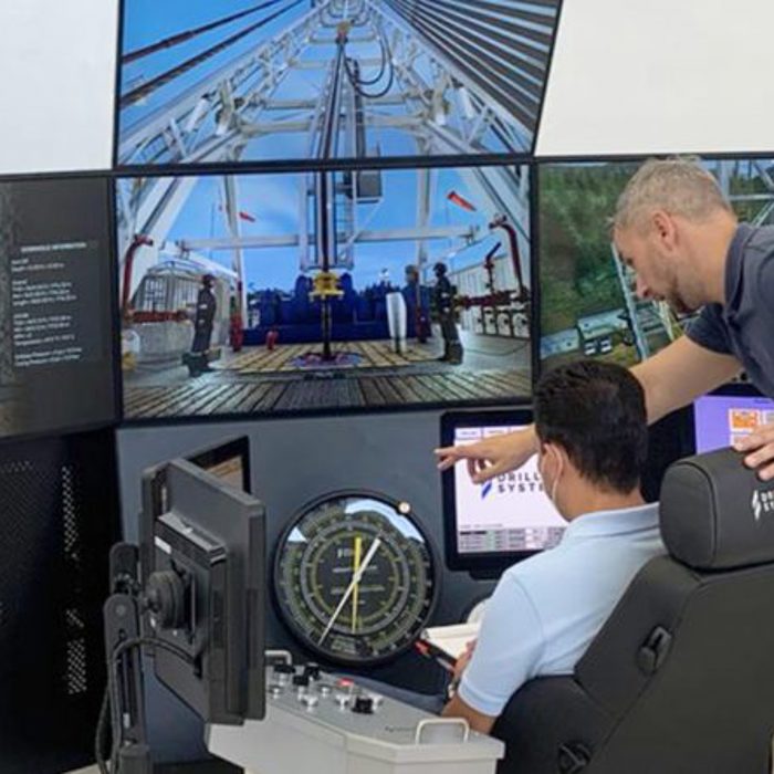 Two individuals operate a high-tech control station with multiple monitors showing industrial equipment and data. One person sits, working the controls, while the other stands, pointing at a screen.