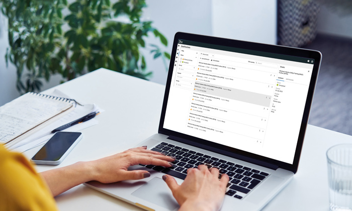 شخص يكتب على جهاز كمبيوتر محمول على مكتب مع صندوق بريد إلكتروني مفتوح على الشاشة، وبجانبه دفتر ملاحظات وقلم وهاتف ذكي وكوب من الماء. توجد نبتة في الخلفية.