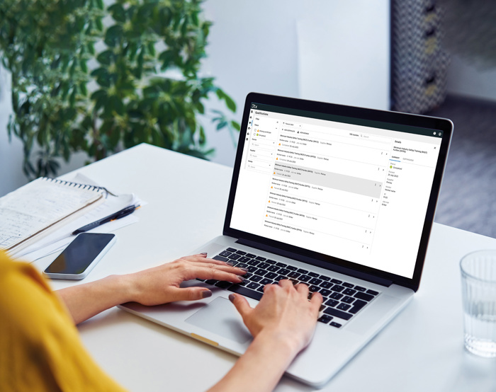 Persona escribiendo en un ordenador portátil en un escritorio con una bandeja de entrada de correo electrónico abierta en la pantalla, junto a un cuaderno, un bolígrafo, un smartphone y un vaso de agua. Al fondo se ve una planta.