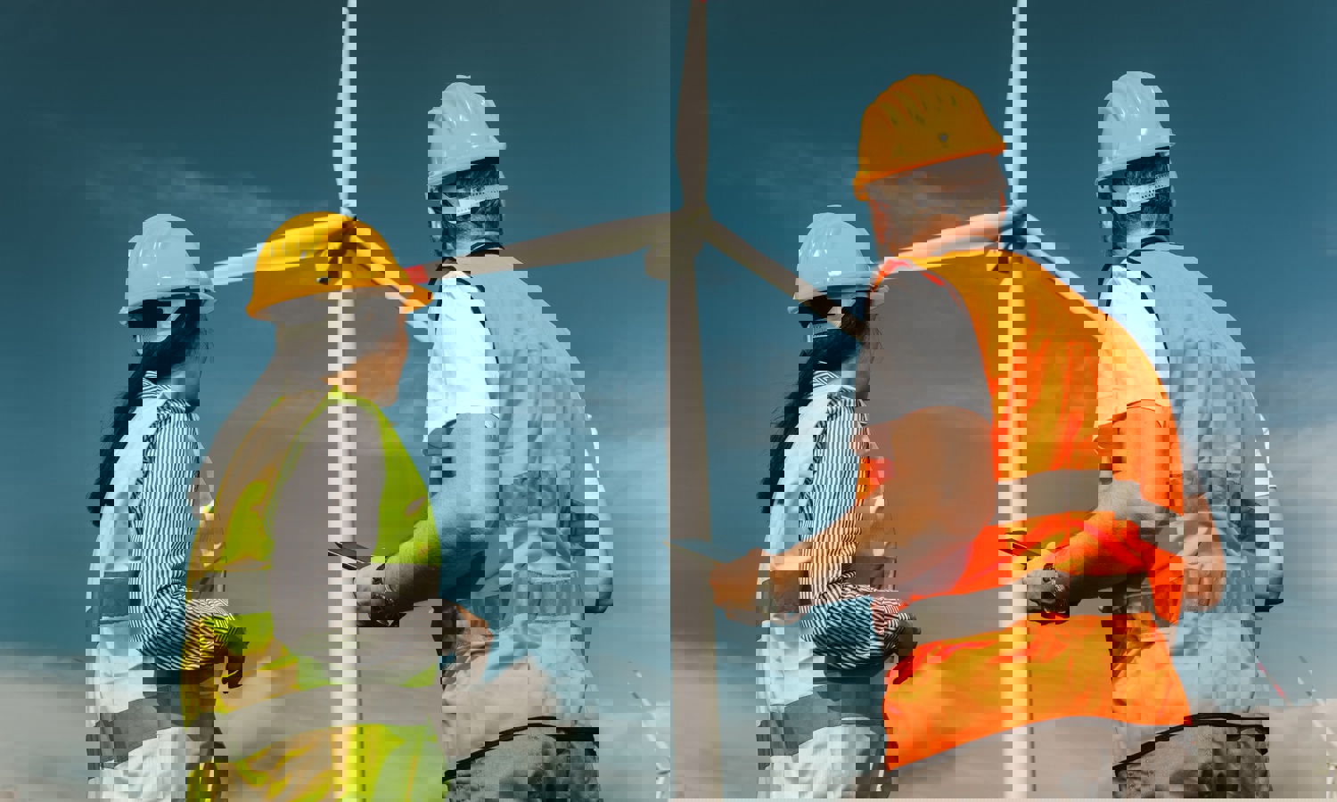 يقف عاملان يرتديان قبعات صلبة وسترات عاكسة للضوء بالقرب من توربين رياح كبير، يراقبان وربما يتفقدانها تحت سماء زرقاء صافية.