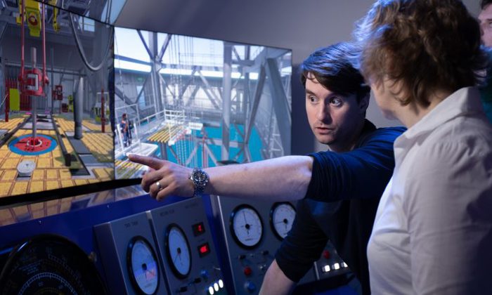 شخص يشير إلى شاشة تعرض أدوات التحكم الصناعية ومحاكاة الآلات أثناء الشرح لشخص آخر أمام لوحات تحكم ذات مقاييس تناظرية.