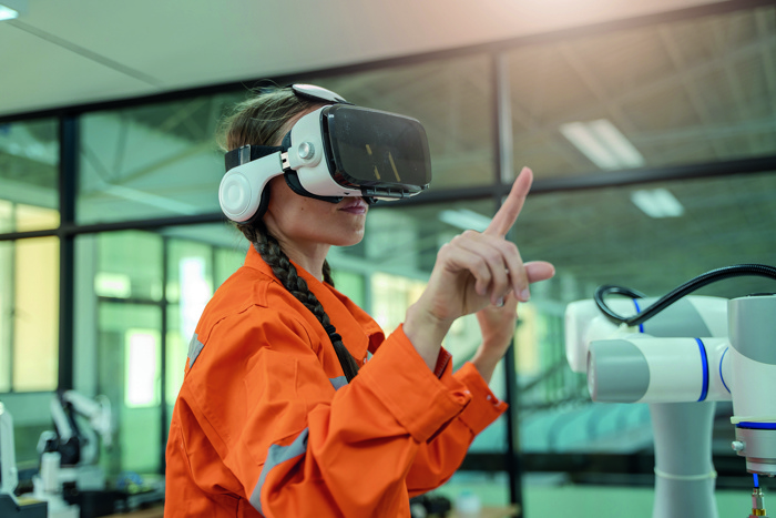 Una persona con una chaqueta naranja y un casco de realidad virtual interactúa con un dispositivo robótico en un moderno entorno industrial.