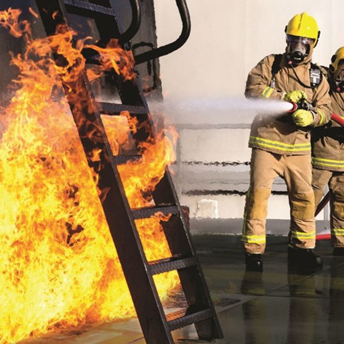 Two firefighters in full gear, including helmets and breathing apparatus, extinguishing a large blaze with a hose on an outdoor metal structure.