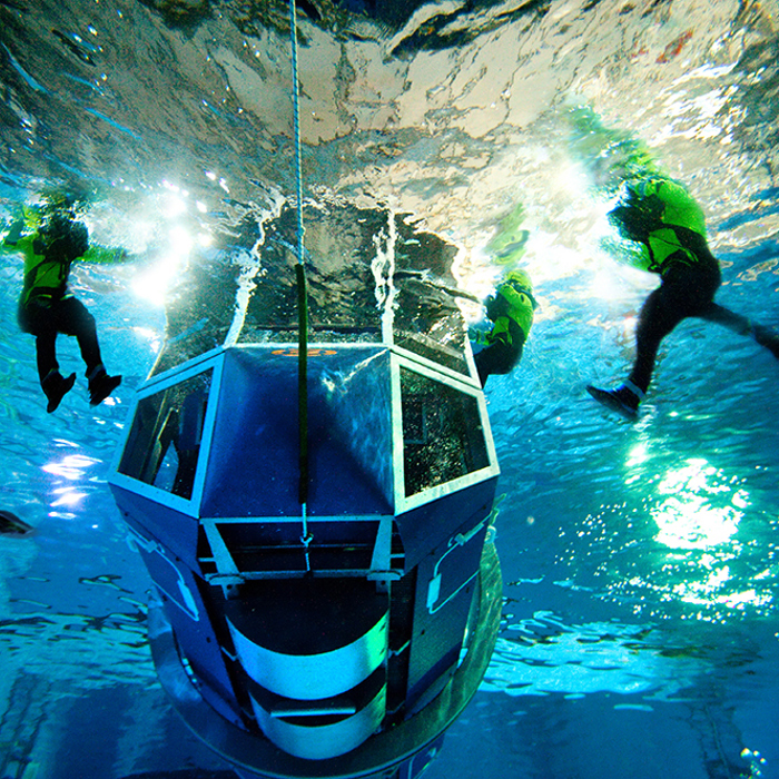 Several divers in scuba gear surround a submerged, large, windowed structure underwater, illuminated by bright lights.