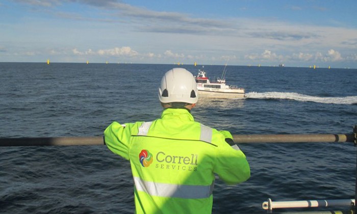 Uma pessoa com um casaco de alta visibilidade e um capacete observa um barco no oceano. O casaco tem o logótipo "Correll Services" nas costas.