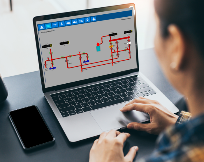 Uma pessoa utiliza um computador portátil que apresenta um diagrama esquemático de um sistema técnico com tubagens e instrumentos. Um smartphone e uma chávena estão sobre a secretária.