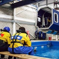 Um grupo de pessoas com equipamento de segurança amarelo e azul senta-se em bancos, observando um cockpit de helicóptero simulado suspenso por cima de uma piscina numa instalação de treino interior.