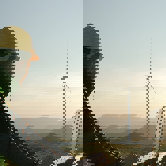 Persona con casco y chaleco de seguridad utilizando un ordenador portátil al aire libre cerca de turbinas eólicas, con un paisaje montañoso de fondo durante la puesta de sol.