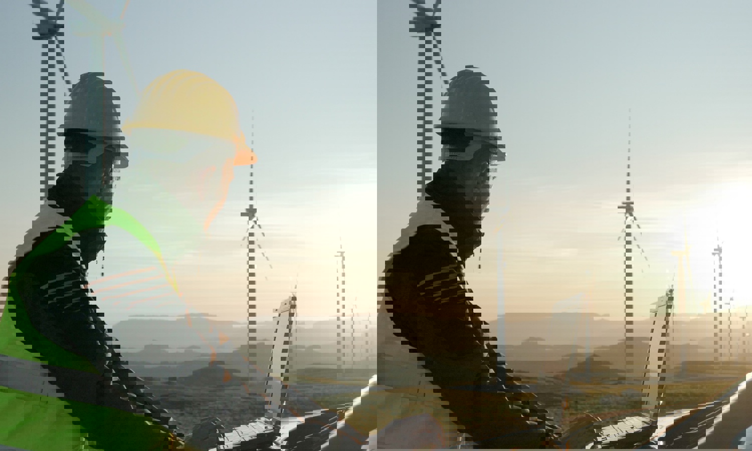 Pessoa com capacete e colete de segurança a utilizar um computador portátil ao ar livre, perto de turbinas eólicas, com uma paisagem montanhosa ao fundo durante o pôr do sol.