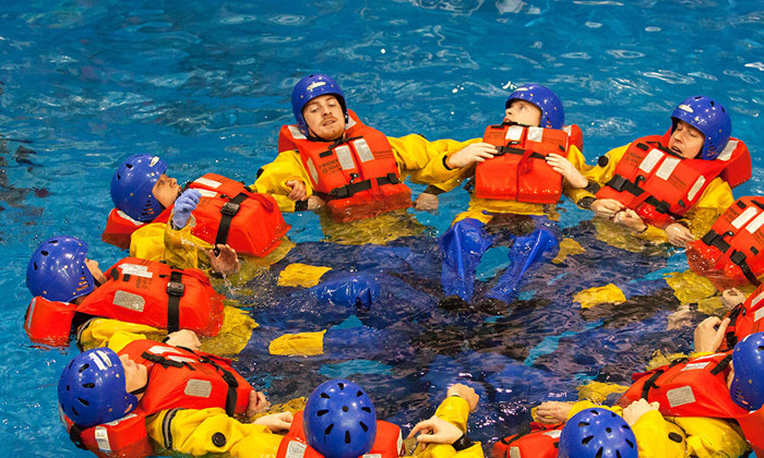 Um grupo de pessoas com coletes salva-vidas e capacetes forma um círculo enquanto flutua numa piscina, assemelhando-se a uma formação militar estratégica. Uma pessoa com um capacete branco observa a zona.