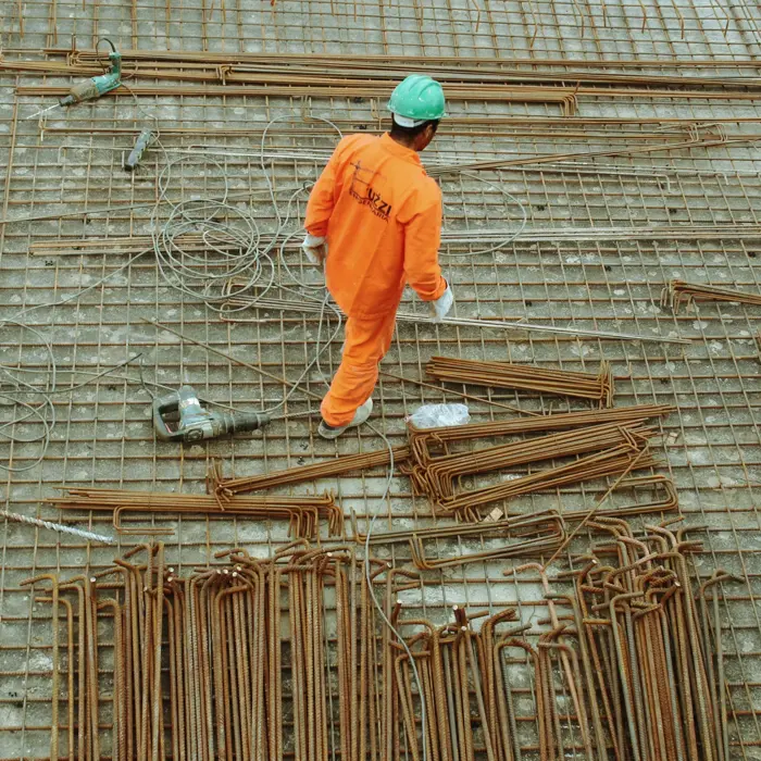 Um trabalhador da construção civil, com um uniforme cor de laranja e um capacete verde, caminha sobre um pavimento de vergalhões e grelha, rodeado de barras e cabos metálicos.