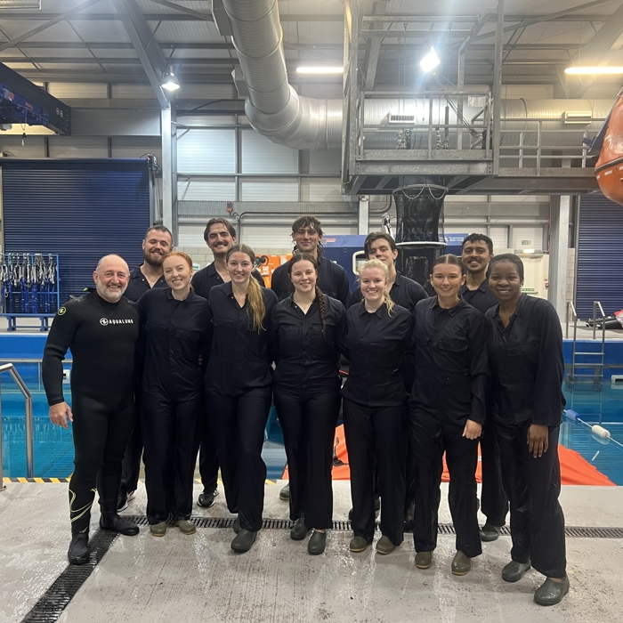 A group of people in black outfits stands together in an indoor facility near a pool and safety equipment, posing for a group photo.