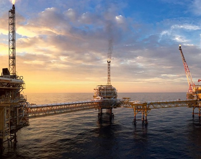Uma plataforma petrolífera offshore ao pôr do sol com uma rede de passadiços ligados sobre o oceano e uma grua do lado direito.
