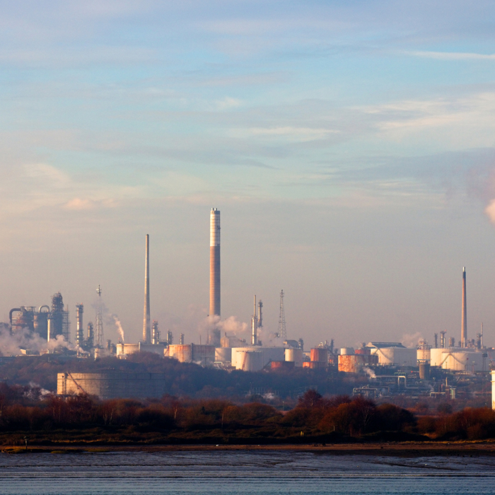 El paisaje de una refinería industrial se extiende bajo un cielo azul con nubes dispersas, sus chimeneas echan humo como centinelas en formación, sincronizadas y firmes.