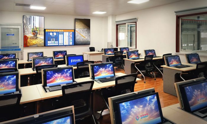 Uma sala de aula moderna equipada com vários postos de trabalho, cada um com um computador que apresenta um protetor de ecrã colorido. Um grande ecrã digital e um quadro branco são visíveis na parte da frente da sala.