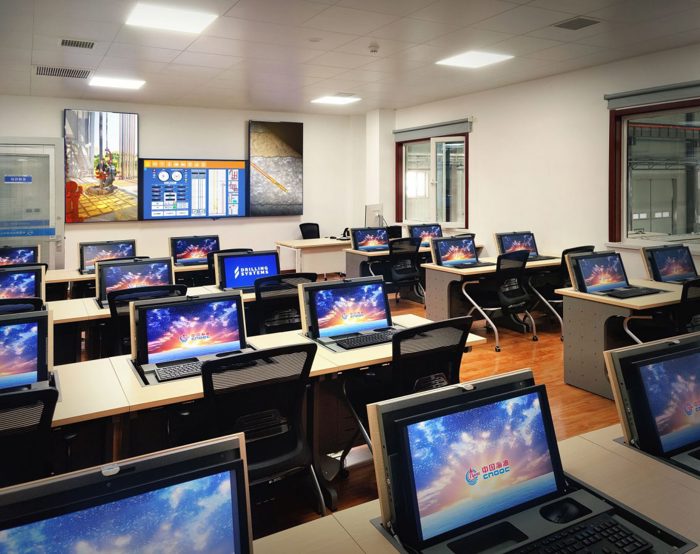 Uma sala de aula moderna equipada com vários postos de trabalho, cada um com um computador que apresenta um protetor de ecrã colorido. Um grande ecrã digital e um quadro branco são visíveis na parte da frente da sala.
