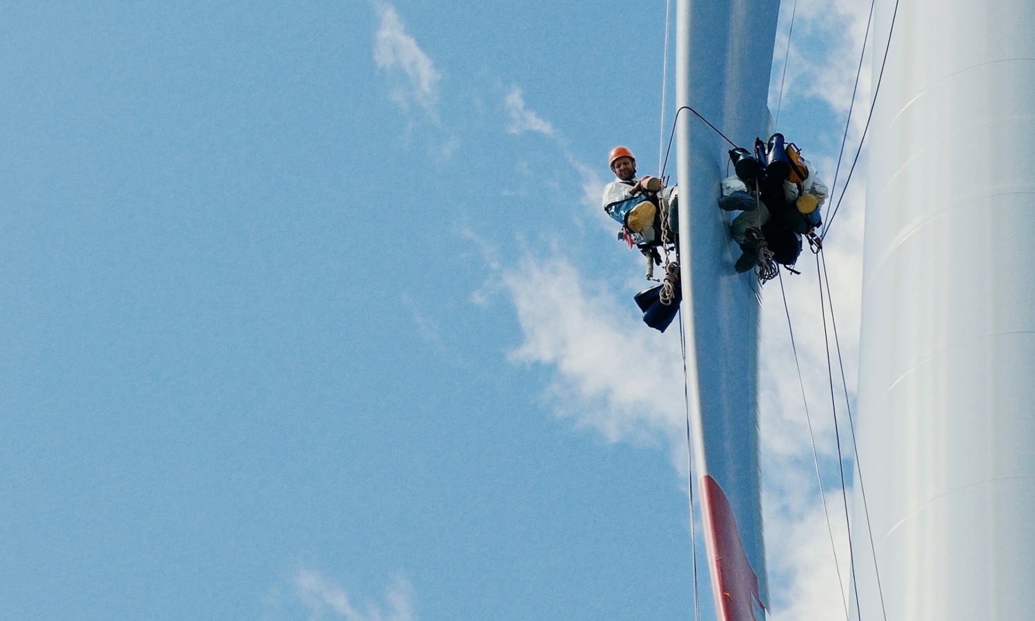 Dos trabajadores con equipo de seguridad, demostrando su experta formación, están suspendidos por cuerdas junto a un aerogenerador contra un cielo azul.