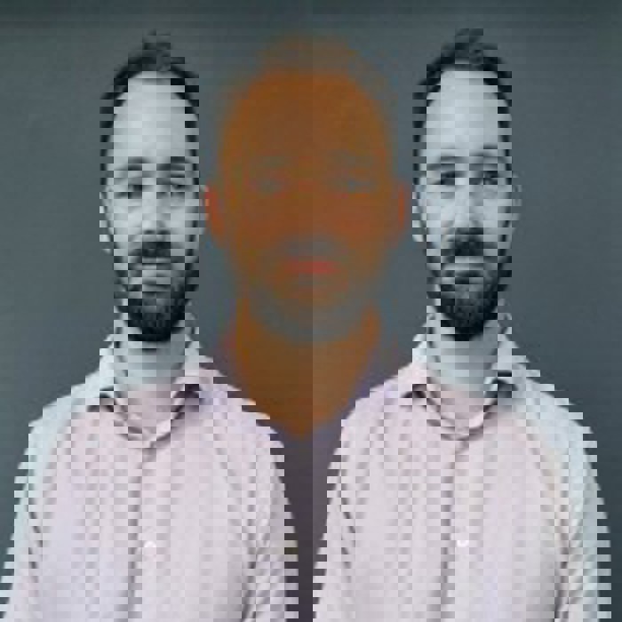 Homem com cabelo curto e barba, vestindo uma camisa de cor clara, de pé, confiante, como se estivesse pronto para treinar, num fundo escuro liso.