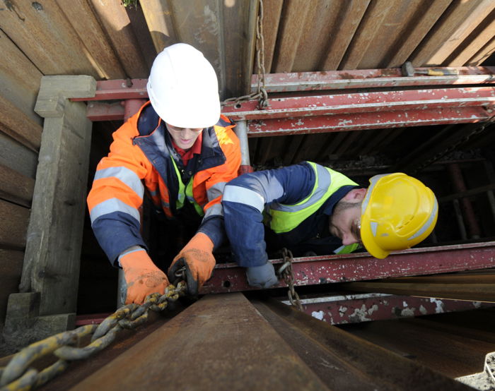 Dos trabajadores de la construcción con equipo de seguridad manipulan una cadena metálica dentro de una zanja con soportes de madera y metal alrededor, aplicando hábilmente su formación para garantizar un proceso seguro y eficaz.