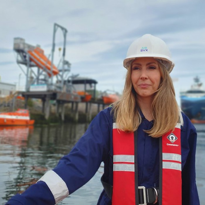 A woman in a hard hat and life jacket stands by the waterfront with boats and industrial equipment in the background.