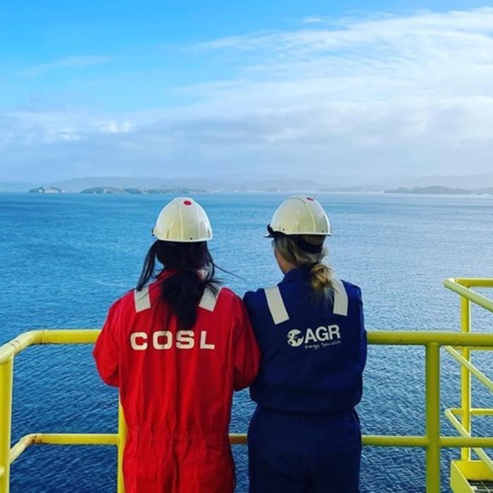 Two workers in helmets and coveralls, labeled COSL and AGR, stand on an offshore platform railing, overlooking a vast expanse of blue sea and distant land.